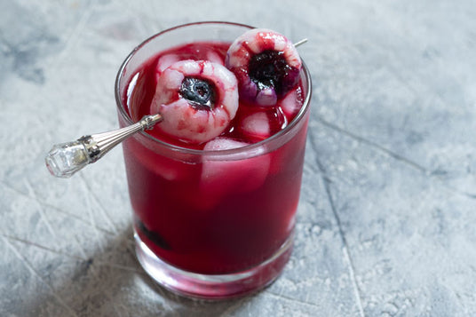 Ein Glas mit rotem Getränk, Eiswürfeln und zwei mit Heidelbeeren gefüllten Litschis, die auf einem Cocktailspieß aufgespießt sind, auf einer grauen Unterlage.