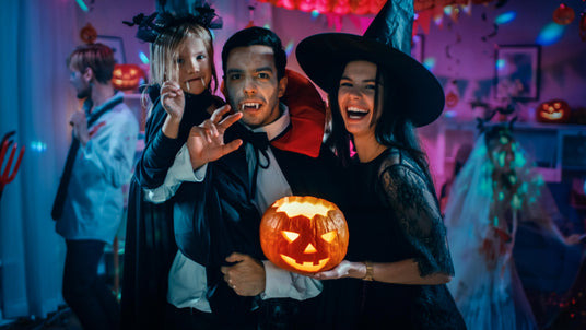 Drei Personen in Halloween-Kostümen, darunter ein Vampir und eine Hexe, stehen lächelnd mit einer geschnitzten Kürbislaterne in einem festlich geschmückten Raum.