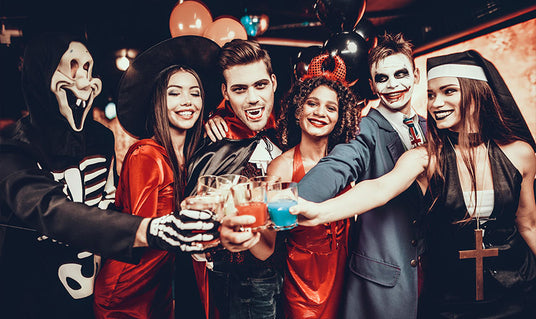 Sechs Personen in Halloween-Kostümen – darunter ein Skelett, eine Hexe, ein Vampir, ein Teufel, ein Clown und eine Nonne – stehen gemeinsam in einem Raum, heben bunte Getränke in die Höhe und lächeln in die Kamera.