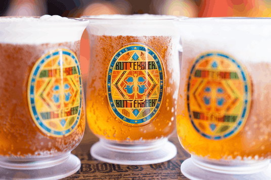 Drei frostige Plastikbecher, gefüllt mit goldenem Butterbier und jeweils mit einem bunten Etikett versehen, stehen nebeneinander vor einem verschwommenen Hintergrund.