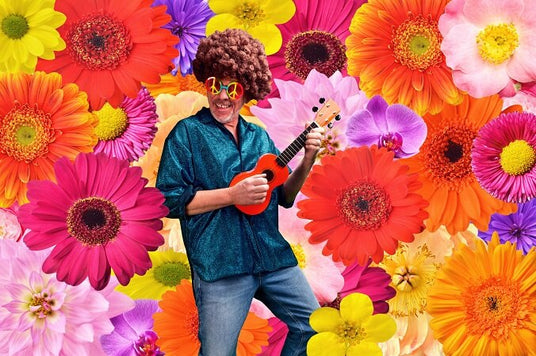 Ein Mann in einem glänzenden Hemd und einer Clownperücke spielt auf einer kleinen Gitarre vor einem farbenfrohen Hintergrund aus großen, leuchtenden Blumen.