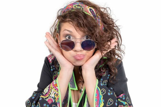 Eine Frau mit lockigem Haar, die eine runde Sonnenbrille und ein buntes Stirnband trägt, macht eine verspielte Grimasse und drückt dabei mit den Händen ihre Wangen zusammen. Sie trägt eine gemusterte Jacke. Weißer Hintergrund.