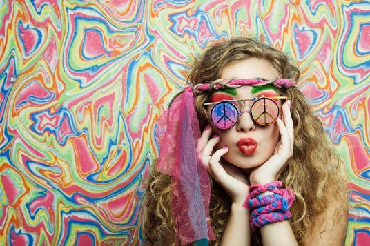 Eine Person mit lockigem Haar trägt eine Brille mit Peace-Zeichen, ein geflochtenes Stirnband und bunte Accessoires und posiert vor einem lebhaften, psychedelischen Hintergrund.