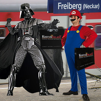 Zwei Personen in Darth-Vader- und Mario-Kostümen halten Einkaufstüten vor einem Bahnhofsschild mit der Aufschrift „Freiberg (Neckar)“, im Hintergrund ist ein roter Zug zu sehen.