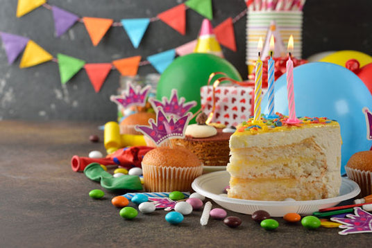 Ein Stück Geburtstagskuchen mit Kerzen, Cupcakes, Luftballons, bunten Süßigkeiten und Partydekoration auf einem Tisch. Im Hintergrund hängen dreieckige Wimpelketten.