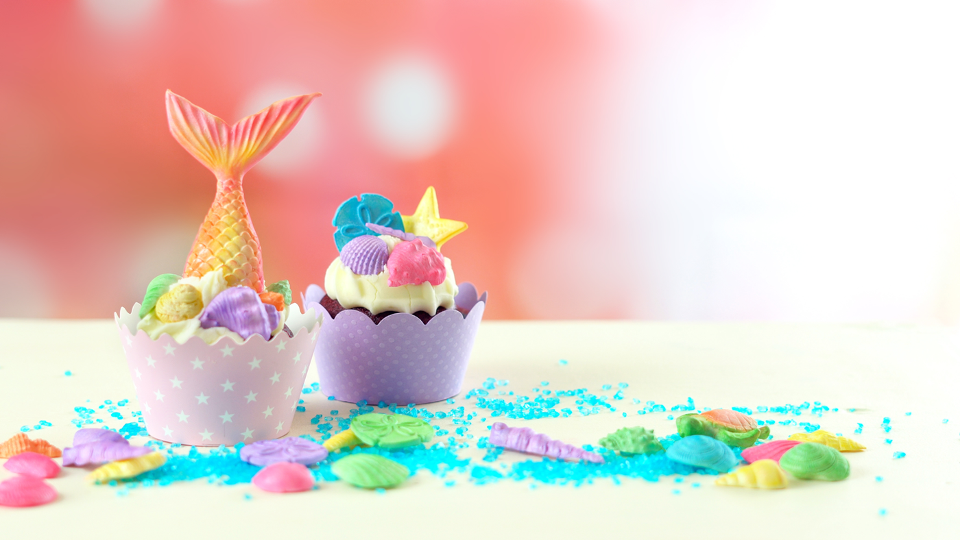 Zwei verzierte Cupcakes mit Meerjungfrauen-Schwänzen und Süßigkeiten zum Thema Meer stehen auf einer weißen Fläche, die mit bunten Streuseln bestreut ist, vor einem rosa, verschwommenen Hintergrund.