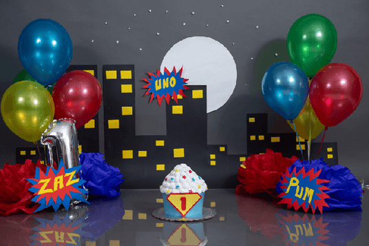 Dekoration zum ersten Geburtstag im Superhelden-Stil mit Luftballons, einem Luftballon in Form der Zahl 1, einer Kulisse mit Stadtansicht und einer Cupcake-Torte mit der Zahl 1 darauf.