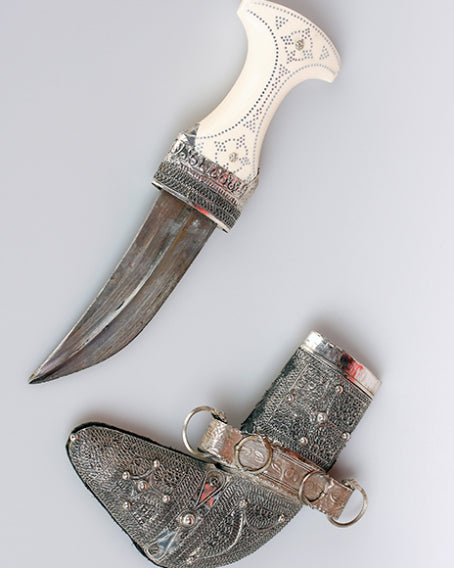 Ein geschwungener Dolch mit einem kunstvoll verzierten weißen Griff und einer verzierten Metallscheide, dargestellt vor einem hellgrauen Hintergrund.