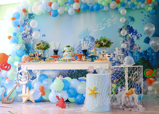 Ein Party-Tisch im Meeresthema mit Meeresdekorationen, Luftballons, ausgeschnittenen Meerestieren und einer Torte vor einem blauen Unterwasser-Hintergrund.