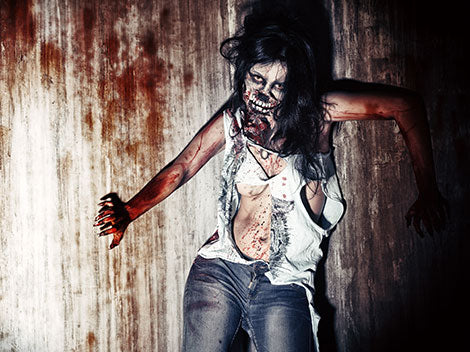 Eine Person in zerlumpten Kleidern und mit Zombie-Make-up steht vor einer fleckigen Wand; sie wirkt zerzaust und bedrohlich, mit Blut und künstlichen Wunden im Gesicht und am Körper.