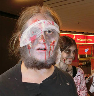 Eine Person mit einem Verband um den Kopf und Gesichts-Make-up, das Wunden und Blut nachahmt, ist als Zombie verkleidet; hinter ihr steht eine weitere Person mit Zombie-Make-up. Sie scheinen sich in einem öffentlichen Innenraum zu befinden.