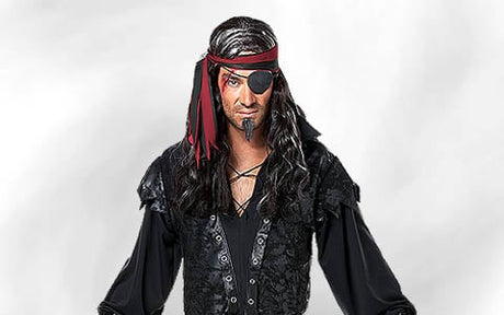 Eine Person in einem schwarzen Piratenkostüm mit einem rot-schwarzen Stirnband, einer Augenklappe, einer langen schwarzen Perücke und einem künstlichen Ziegenbart, die vor einem schlichten hellen Hintergrund steht.
