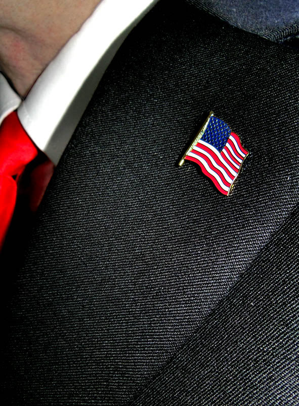Nahaufnahme einer Person, die einen schwarzen Anzug, eine rote Krawatte und eine USA-Flaggen-Anstecknadel – eine elegante Anstecknadel mit der amerikanischen Flagge – am Revers trägt.