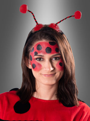 Eine Person in einem Marienkäferkostüm mit Gesichtsbemalung, einem rot-schwarz gepunkteten Käfer-Outfit und einem Stirnband mit Marienkäfer-Fühlern und roten Pompons vor einem grauen Hintergrund.