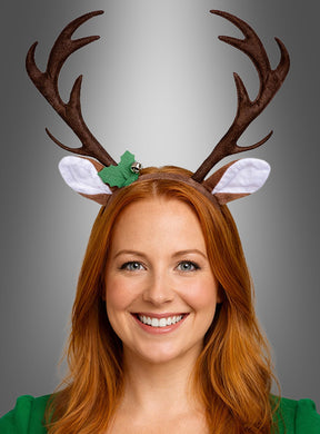 Eine lächelnde Frau mit roten Haaren, die ein „Rentier-Geweih mit Ohren und Glöckchen“ trägt, das aus einem braunen Geweih, weißen Ohren, einem grünen Blatt und einem kleinen Glöckchen besteht. Das verspielte Accessoire rundet ihren bezaubernden Reh-Kostüm-Look ab.