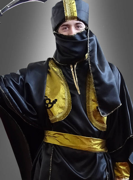 Eine Person, gekleidet in ein schwarz-goldenes Tuareg-Orientkostüm „Ölscheich 1001 Nacht“ und mit einer Kopfbedeckung, bei der nur die Augen und der obere Teil der Nase zu sehen sind, posiert vor einem schlichten grauen Hintergrund.