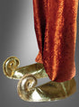 Eine Person in einer weiten orangefarbenen Hose und glänzenden goldenen „Wüstenprinz“-Schnabelschuh-Überzügen mit nach oben gebogenen Zehen, die an „1001 Nacht“ erinnern, steht vor einem neutralen Hintergrund.