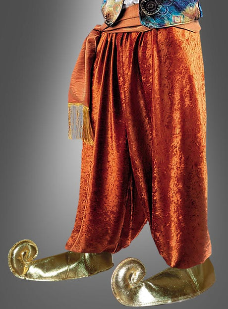 Person, die eine weite, orangefarbene Samthose mit einer Schärpe und goldene „Wüstenprinz“-Schnabelschuh-Überzieher mit nach oben gebogenen Zehen trägt, die an „1001 Nacht“ erinnern. Der Oberkörper ist teilweise zu sehen. Neutraler Hintergrund mit Farbverlauf.