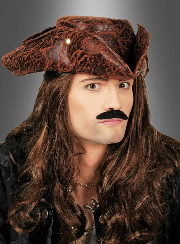 Eine Person, die einen braunen Piratenhut (Dreispitz in Lederoptik), eine lange braune Perücke, einen falschen Schnurrbart, eine schwarze Jacke und eine Halskette trägt, posiert vor einem grauen Hintergrund mit Farbverlauf.