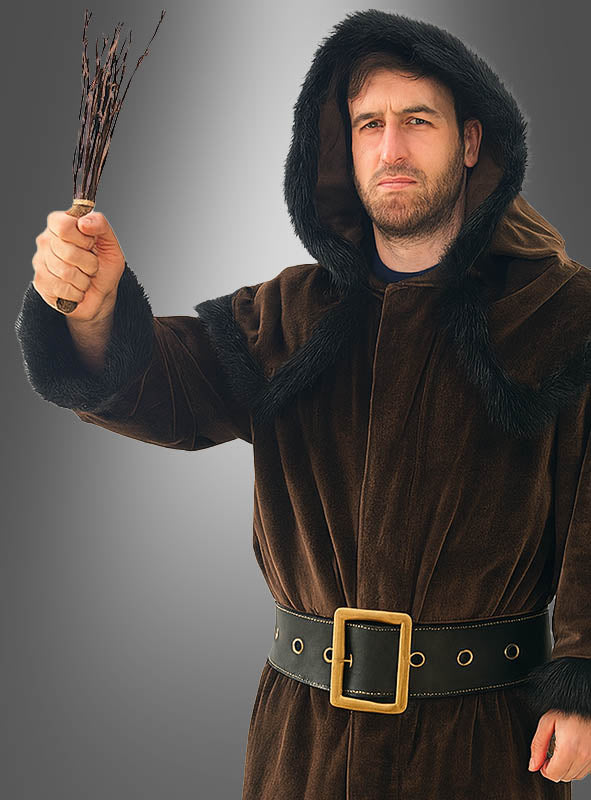 Ein Mann, der das Knecht-Ruprecht-Kostüm – einen braunen Mantel mit Kapuze und schwarzem Pelzbesatz – trägt, hält ein kleines Bündel Zweige in der Hand und blickt ernst.