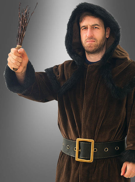 Ein Mann, der das Knecht-Ruprecht-Kostüm – einen braunen Mantel mit Kapuze und schwarzem Pelzbesatz – trägt, hält ein kleines Bündel Zweige in der Hand und blickt ernst.
