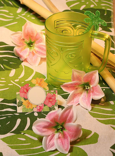 Ein grüner Tiki-Becher, rosa Kunstblumen, Holzstäbchen und eine Karte mit Blumenmotiv sind auf dem „Bedruckter Palmblätter Tischläufer 3 Meter“ arrangiert – einem grünen Tischläufer mit Blattmuster und eleganten Monstera-Blättern –, der sich perfekt als Partydekoration oder als auffälliger Hawaii-Tischläufer für Ihre nächste tropische Feier eignet.
