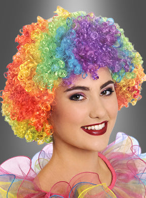 Eine Person mit einer Regenbogen-Afro-Perücke und auffälligem Make-up, lächelnd, mit einem gerüschten, bunten Kostümkragen vor einem grauen Hintergrund.