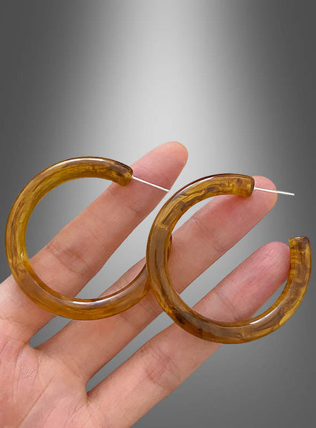 Eine Hand hält ein Paar klobige, durchscheinende braune Hippie-Creolen vor einem grauen Hintergrund mit Farbverlauf.