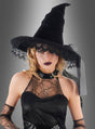 Eine Frau in einer edlen Hexenhütte aus Samt mit Spitze und einem Spinnennetzkleid, die dunklen Lippenstift trägt, posiert vor einem grauen Hintergrund – ein auffälliges Halloween-Kostüm, das perfekt zur Jahreszeit passt.