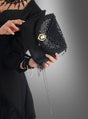 Eine Person in schwarzer Kleidung hält die schwarze Gothic-Handtasche mit Brosche vor einem grauen Hintergrund mit Farbverlauf in der Hand; die Tasche ist mit Pailletten, Kettendetails und einer Brosche im Kameen-Stil verziert.