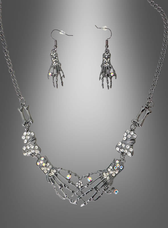 Ein „Skelett Hand“-Schmuckset aus Silber, bestehend aus einer Halskette und Ohrringen in Form von Skeletthänden, die mit klaren Strasssteinen verziert sind, wird vor einem grauen Hintergrund mit Farbverlauf präsentiert.