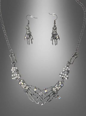 Ein „Skelett Hand“-Schmuckset aus Silber, bestehend aus einer Halskette und Ohrringen in Form von Skeletthänden, die mit klaren Strasssteinen verziert sind, wird vor einem grauen Hintergrund mit Farbverlauf präsentiert.