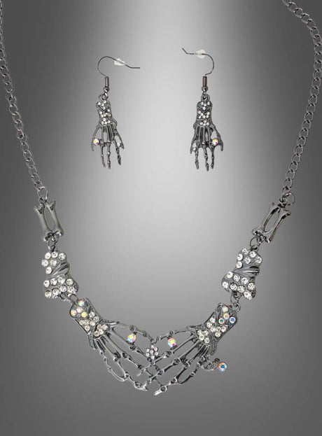 Ein „Skelett Hand“-Schmuckset aus Silber, bestehend aus einer Halskette und Ohrringen in Form von Skeletthänden, die mit klaren Strasssteinen verziert sind, wird vor einem grauen Hintergrund mit Farbverlauf präsentiert.