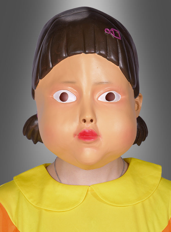 Eine Person, die die „Game Puppe Maske koreanisches Mädchen“ trägt – eine große Plastikmaske, die einem karikaturhaften Gesicht mit großen Augen und rosa Lippen ähnelt – und dazu ein gelbes Outfit trägt, steht vor einem grauen Hintergrund mit Farbverlauf.