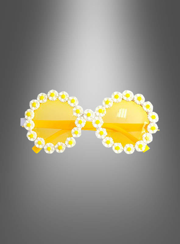 Vor einem grauen Hintergrund mit Farbverlauf präsentiert sich die Hippie-Blumenbrille „Gänseblümchen“ mit einem runden, gelben Oversize-Rahmen, der mit weißen und gelben Kunstgänseblümchen verziert ist.