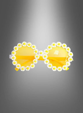 Vor einem grauen Hintergrund mit Farbverlauf präsentiert sich die Hippie-Blumenbrille „Gänseblümchen“ mit einem runden, gelben Oversize-Rahmen, der mit weißen und gelben Kunstgänseblümchen verziert ist.