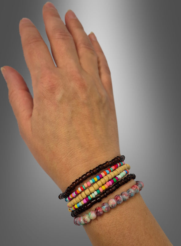 Eine linke Hand, an der vier braun-bunte „Hippie Perlen“-Armbänder aus einem Set getragen werden, die an Schmuck aus den 70er Jahren erinnern, vor einem grauen Hintergrund mit Farbverlauf.