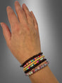 Eine linke Hand, an der vier braun-bunte „Hippie Perlen“-Armbänder aus einem Set getragen werden, die an Schmuck aus den 70er Jahren erinnern, vor einem grauen Hintergrund mit Farbverlauf.