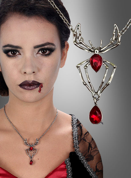 Eine Frau mit dramatischem Make-up und Kunstblut auf der Lippe trägt eine auffällige Spinnen-Halskette mit roten Edelsteinen – eine ins Auge fallende silberne Halskette in Spinnenform, die mit zwei großen roten Edelsteinen verziert ist. Die Halskette eignet sich perfekt als Halloween-Schmuck und ist rechts in einer Nahaufnahme zu sehen.
