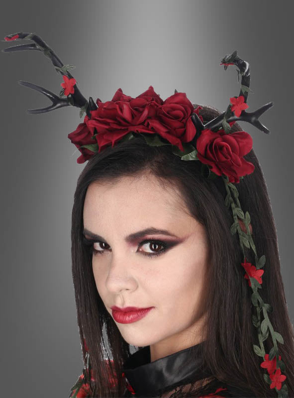 Eine Frau mit auffälligem Make-up trägt das Haarreif „Deluxe Geweih Haarschmuck mit Rosen“, das mit markanten schwarzen Geweihen, leuchtend roten Rosen und üppigen grünen Ranken verziert ist – eine bezaubernde Verschmelzung von Rosen-Kopfschmuck und Dark-Mori-Accessoires – vor einem grau abgestuften Hintergrund.