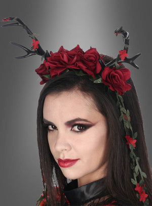 Eine Frau mit auffälligem Make-up trägt das Haarreif „Deluxe Geweih Haarschmuck mit Rosen“, das mit markanten schwarzen Geweihen, leuchtend roten Rosen und üppigen grünen Ranken verziert ist – eine bezaubernde Verschmelzung von Rosen-Kopfschmuck und Dark-Mori-Accessoires – vor einem grau abgestuften Hintergrund.