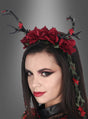 Eine Frau mit auffälligem Make-up trägt das Haarreif „Deluxe Geweih Haarschmuck mit Rosen“, das mit markanten schwarzen Geweihen, leuchtend roten Rosen und üppigen grünen Ranken verziert ist – eine bezaubernde Verschmelzung von Rosen-Kopfschmuck und Dark-Mori-Accessoires – vor einem grau abgestuften Hintergrund.