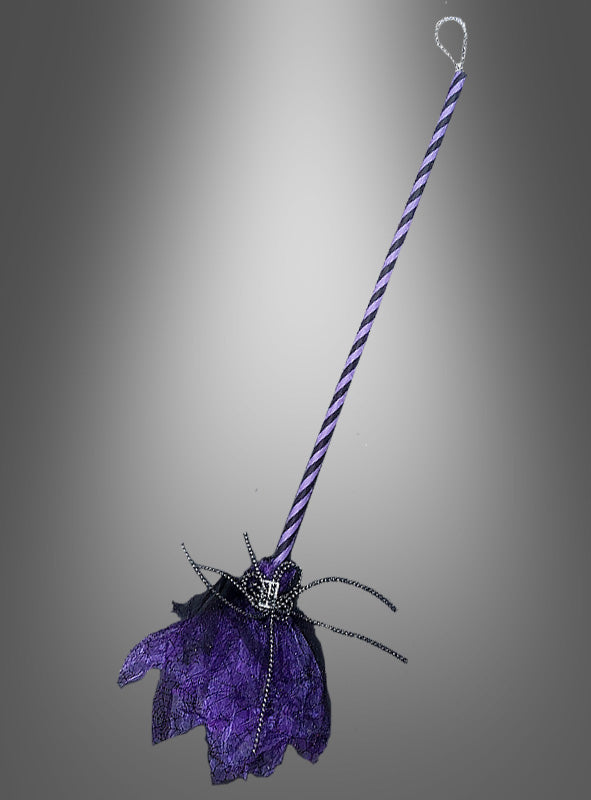 Ein 100 cm langer, schwarz-lila Hexenbesen mit gestreiftem Stiel, borstenartigen Zinken im Spitzenstil und Perlenverzierungen – ideal als Accessoire für ein Hexenkostüm oder als bezaubernde Ergänzung für Ihre Halloween-Party – vor einem grauen Hintergrund mit Farbverlauf.
