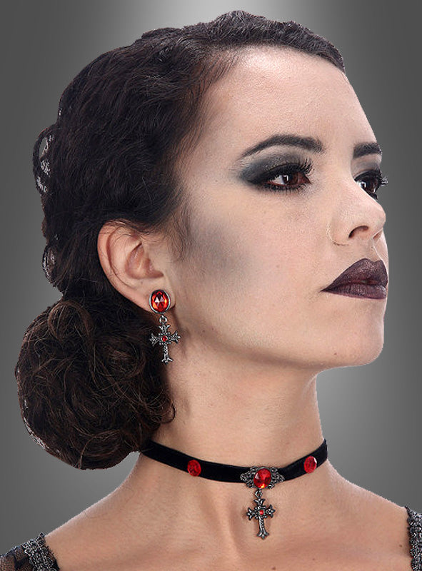 Eine Frau mit dunklem Haar, das zu einem tief sitzenden Dutt zusammengebunden ist, mit Gothic-Make-up und dem Halloween-Schmuckset „Kreuze mit roten Edelsteinen“ – einem Gothic-Schmuckset bestehend aus einem schwarzen Samt-Halsband mit einem Kreuzanhänger, der mit roten Edelsteinen verziert ist, sowie passenden Kreuz-Ohrringen – blickt vor einem grauen Hintergrund zur Seite.