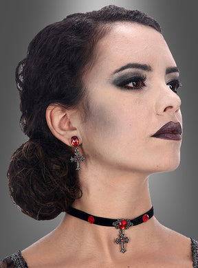 Eine Frau mit dunklem Haar, das zu einem tief sitzenden Dutt zusammengebunden ist, mit Gothic-Make-up und dem Halloween-Schmuckset „Kreuze mit roten Edelsteinen“ – einem Gothic-Schmuckset bestehend aus einem schwarzen Samt-Halsband mit einem Kreuzanhänger, der mit roten Edelsteinen verziert ist, sowie passenden Kreuz-Ohrringen – blickt vor einem grauen Hintergrund zur Seite.