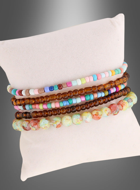 Fünf Perlenarmbänder in Pastelltönen, Braun und bunten Mustern aus dem „Hippie Perlenarmband-Set braun-bunt“ werden auf einem weißen Kissen vor einem grauen Farbverlauf präsentiert und verleihen Ihrem Look mit diesen vom Schmuck der 70er Jahre inspirierten Perlenarmbändern einen Hauch von Retro-Flair.