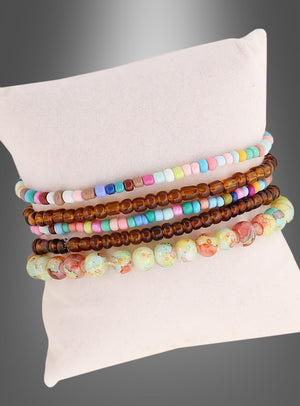 Fünf Perlenarmbänder in Pastelltönen, Braun und bunten Mustern aus dem „Hippie Perlenarmband-Set braun-bunt“ werden auf einem weißen Kissen vor einem grauen Farbverlauf präsentiert und verleihen Ihrem Look mit diesen vom Schmuck der 70er Jahre inspirierten Perlenarmbändern einen Hauch von Retro-Flair.