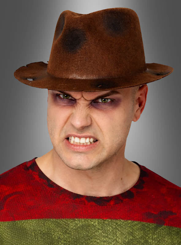 Vor einem grauen Hintergrund steht eine Person, die einen braunen Fedora-Hut der Marke „Freddy“ und ein rot-grünes Hemd trägt, mit zornigem Gesichtsausdruck, zusammengebissenen Zähnen und dunklem Augen-Make-up.