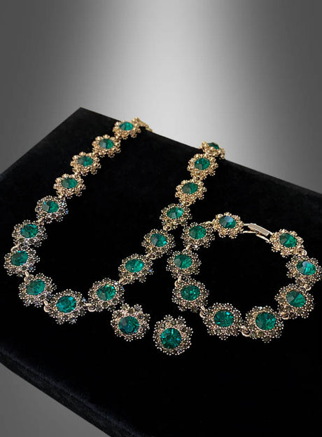 Das „Edles Schmuckset mit Strasssteinen, 4-teilig“ wird auf einer schwarzen Samtunterlage mit grau verlaufendem Hintergrund präsentiert und zeigt seine elegante goldene Halskette und das Armband, die mit großen grünen Edelsteinen und funkelnden Strasssteinen verziert sind – perfekt für jedes Kostüm im Stil der 20er Jahre.