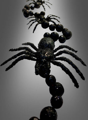 Nahaufnahme der „Schwarze Spinnen Girlande 160 cm“, einer schwarzen, glitzernden Halloween-Dekoration mit Plastikspinnen und Perlen, die in einer Reihe angeordnet sind und vor einem Hintergrund mit Farbverlauf präsentiert werden.
